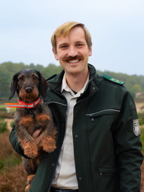 Ein Mann h&auml;lt einen Dackel unter seinem Arm. Er tr&auml;gt eine Bundesforst-Uniform und steht in der Heide.