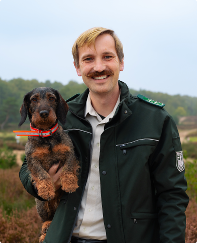 Ein Mann h&auml;lt einen Dackel unter seinem Arm. Er tr&auml;gt eine Bundesforst-Uniform und steht in der Heide.