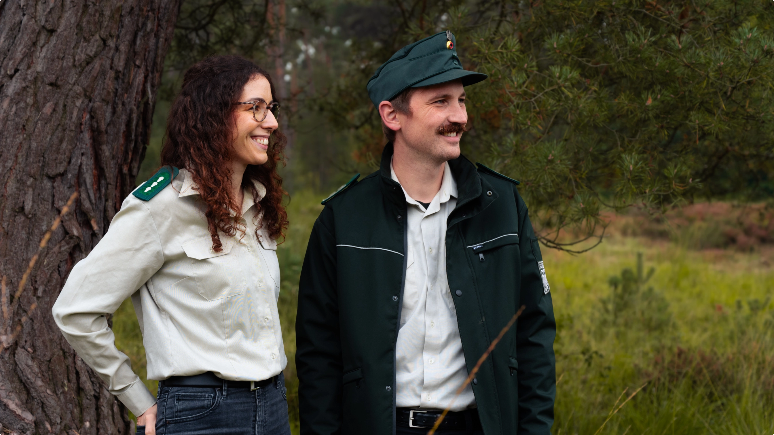 Eine Frau und ein Mann stehen nebeneinander vor einem Baum und blicken lächelnd in die Ferne.